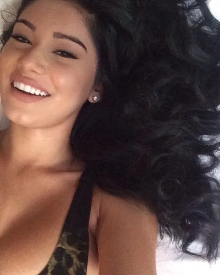 Femme latine aux cheveux bruns, portant un haut noir, regardant vers le côté, avec un fond neutre.