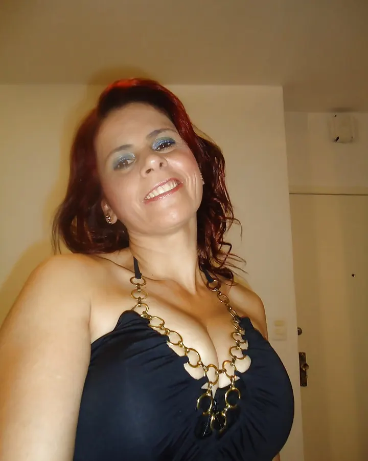 Femme aux cheveux roux longs, portant une robe noire ajustée, debout dans un couloir intérieur avec une main sur la rampe et l'autre sur la hanche.