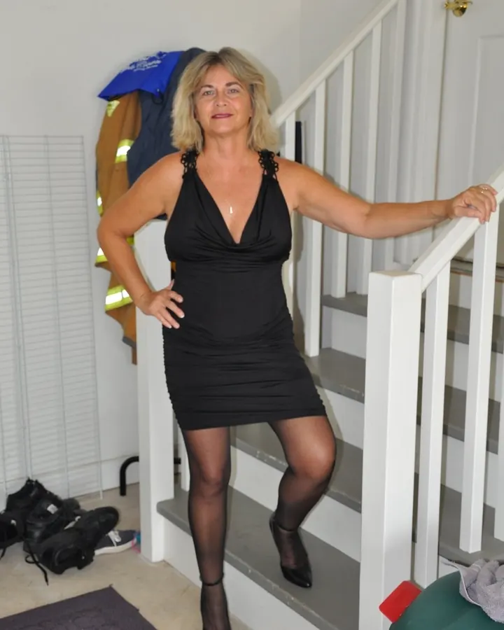 Femme aux cheveux blonds mi-longs, portant une robe noire ajustée, debout sur un escalier intérieur avec une main sur la rampe et l'autre sur la hanche.