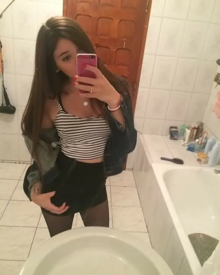 Jeune femme aux cheveux châtains, en haut rayé noir et blanc, prenant un selfie dans une salle de bain.
