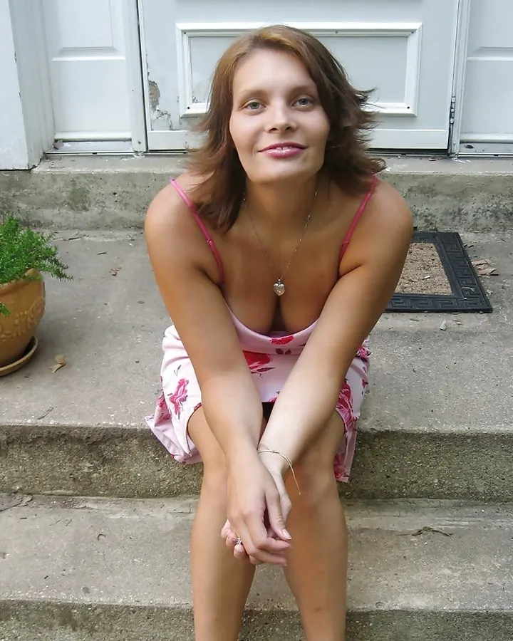 Femme aux cheveux roux en robe rose à fleurs, assise sur des marches devant une porte blanche, souriant doucement.