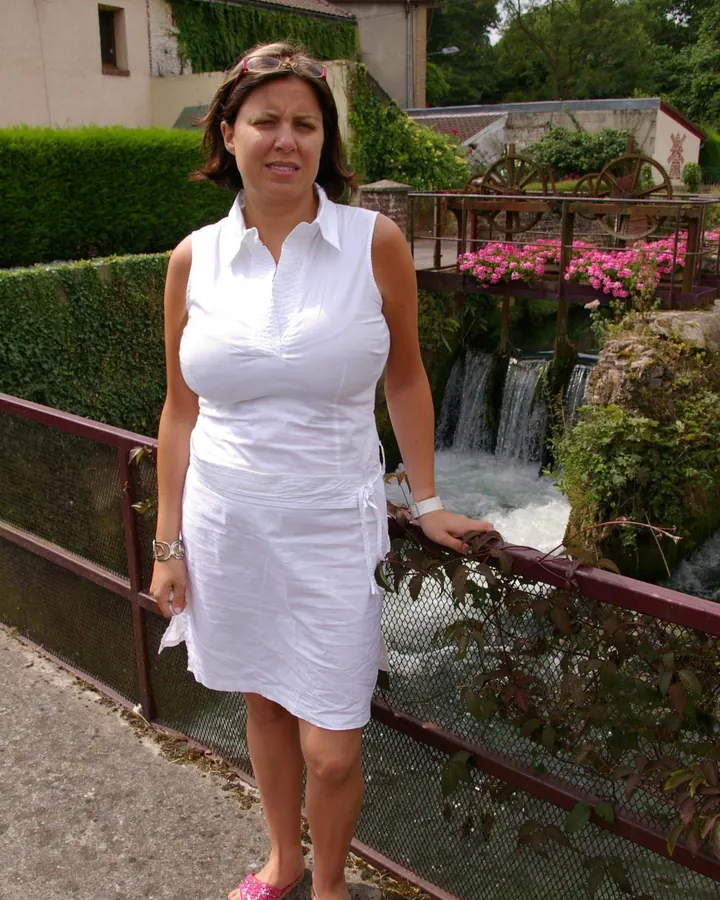 Femme en robe blanche, debout près d'une cascade, avec des fleurs roses en arrière-plan.