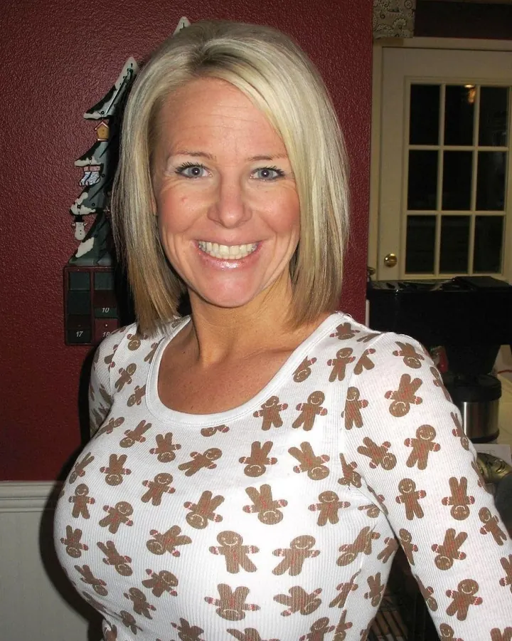 Femme européenne aux cheveux blonds, souriante, vêtue d'un haut noir, posant dans une chambre.