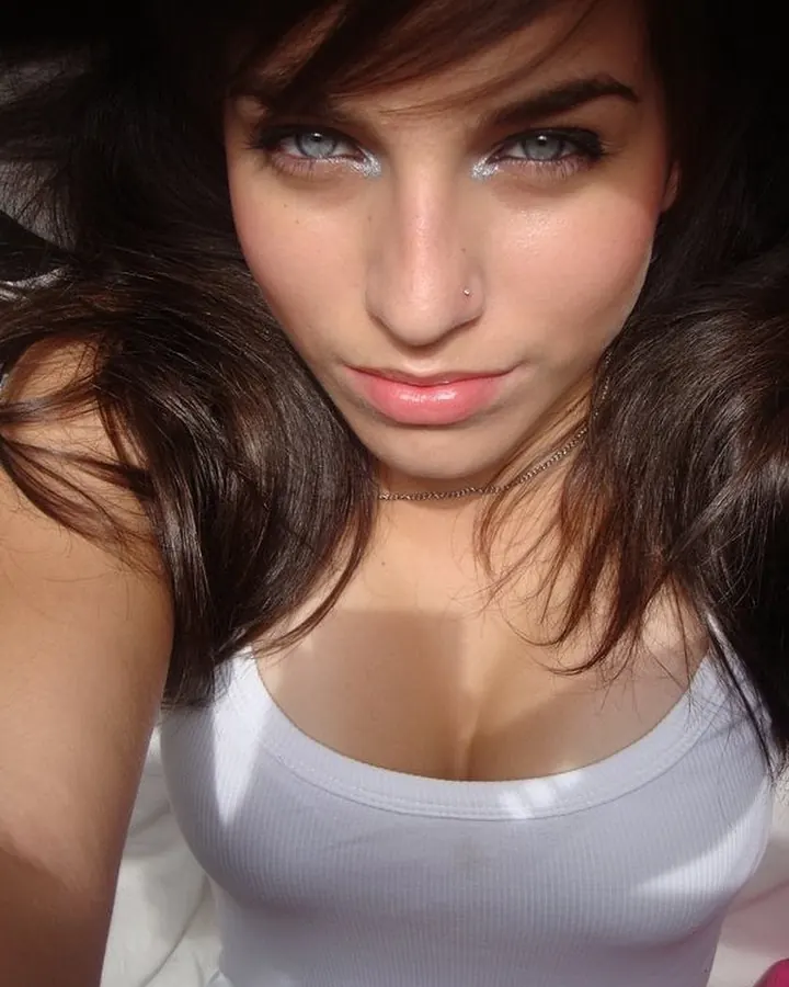 Femme européenne aux cheveux châtains, souriante, vêtue d'un haut blanc, posant dans une pièce sombre.