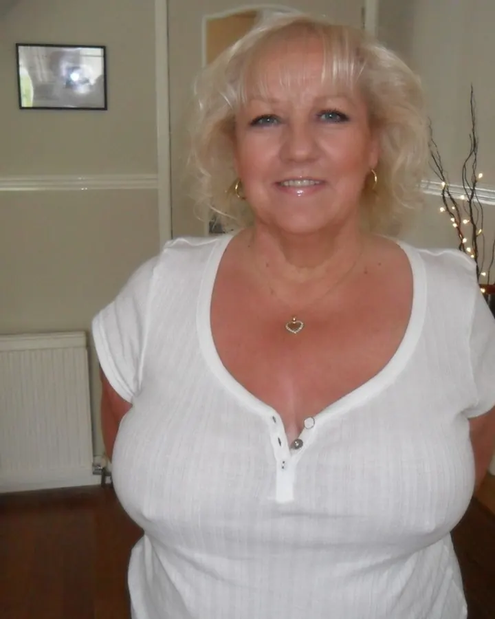 Femme blonde aux cheveux bouclés portant un haut blanc, souriante, dans un intérieur domestique.