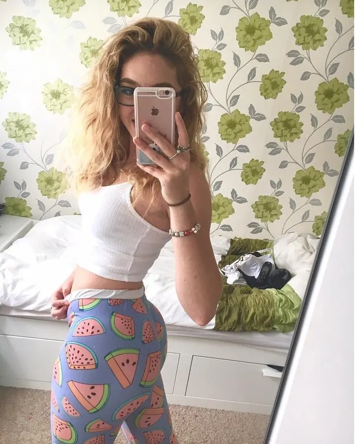 Femme blonde en body rouge, prenant un selfie dans un miroir, dos tourné, dans un studio.