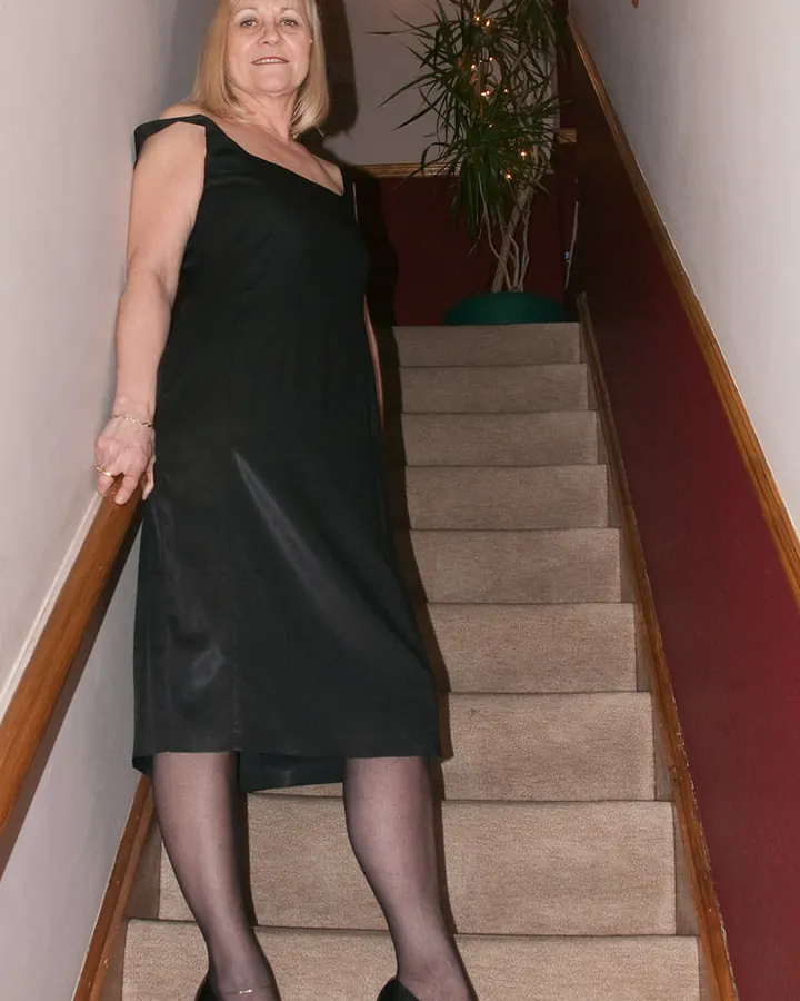 Femme aux cheveux blonds portant une robe noire, descendant un escalier intérieur éclairé par une lampe murale.