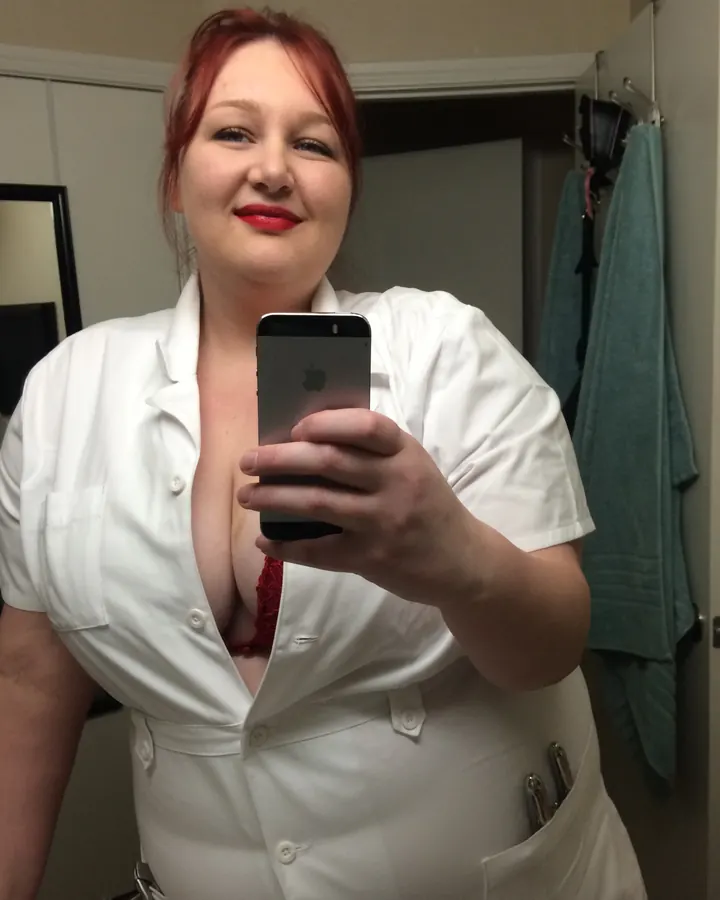 Femme aux cheveux roux portant un haut noir, prenant un selfie dans une salle de bain avec un masque chirurgical bleu.