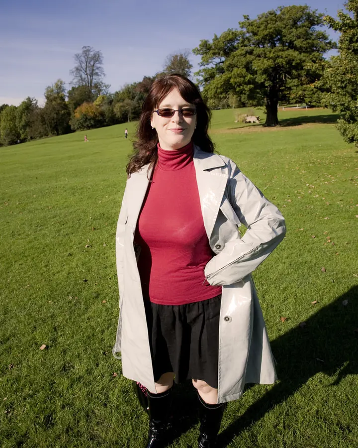 Femme aux cheveux bruns portant une veste grise, un haut rouge et une jupe noire, debout dans un parc ensoleillé.