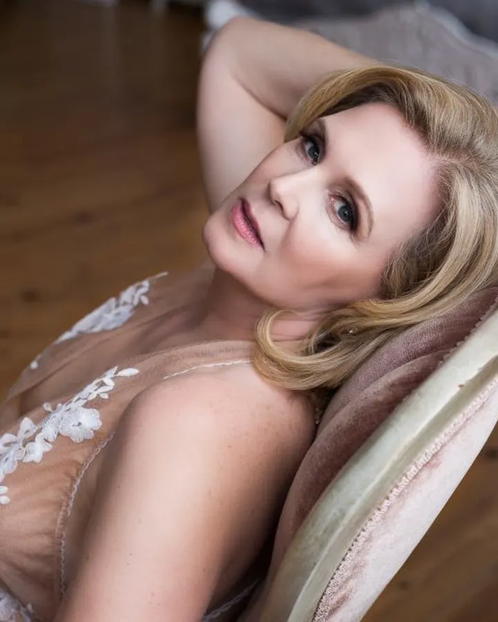 Femme blonde en robe transparente brodée, posant sur un fauteuil, regard vers l'appareil, main dans les cheveux