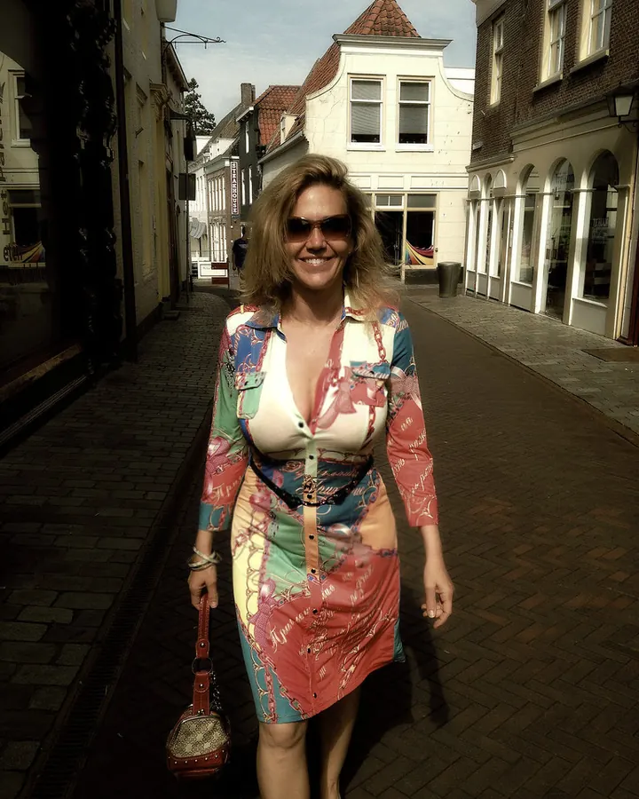 Femme blonde en robe colorée, souriante, posant dans une rue pavée, lunettes de soleil, sac rouge