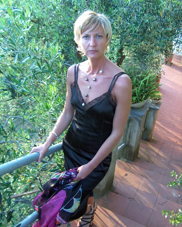 Femme aux cheveux blonds courts, regardant directement l'appareil photo, portant une robe noire en satin, assise sur un balcon avec des plantes en arrière-plan.