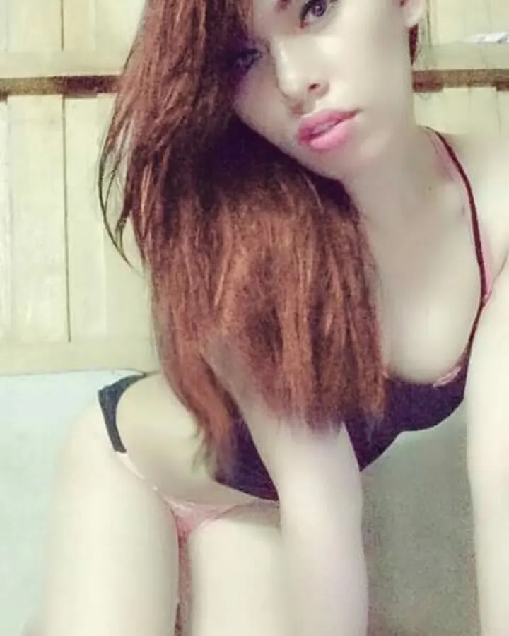 Jeune femme aux cheveux roux, portant un haut noir, un short noir, debout dans une chambre, regardant directement l'appareil photo.