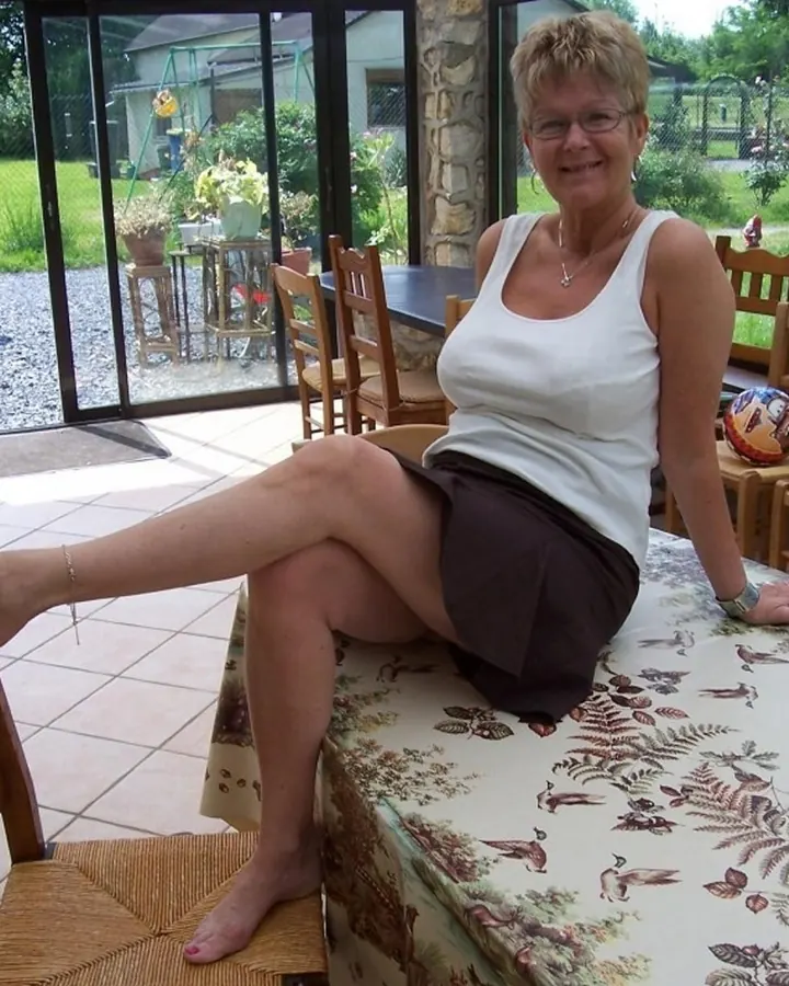 Femme blonde en haut blanc et jupe marron, assise sur une table dans une terrasse, pieds sur une chaise.