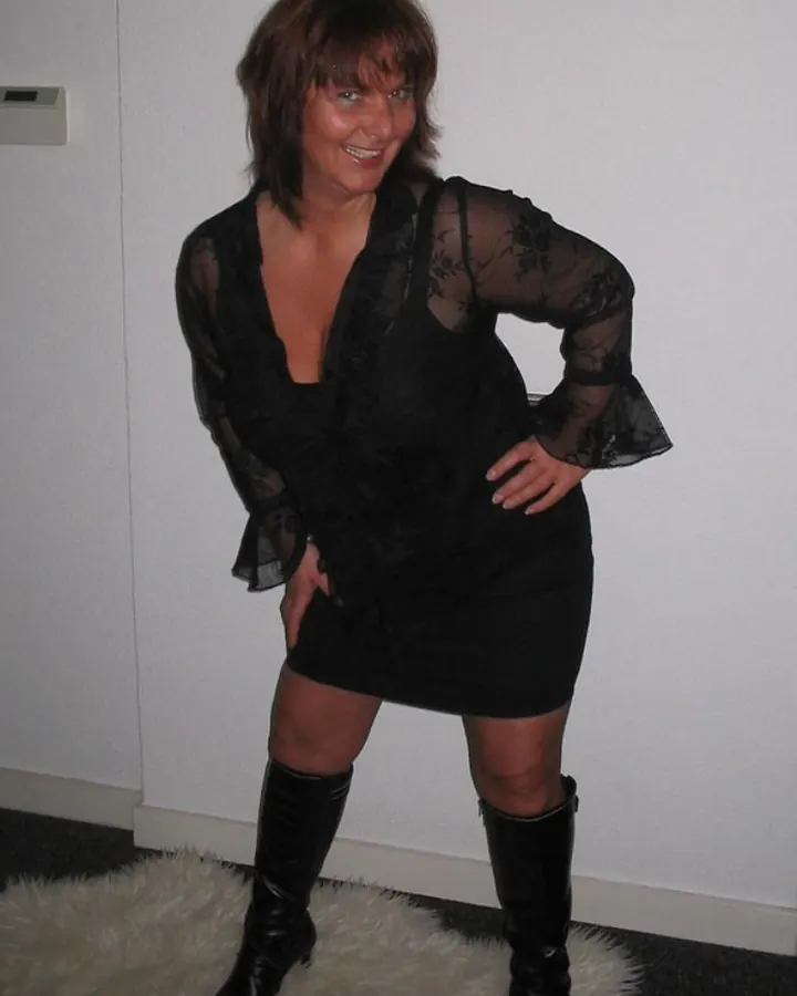 Femme souriante, cheveux bruns, portant une robe noire transparente avec des bottes noires, debout sur un tapis blanc.