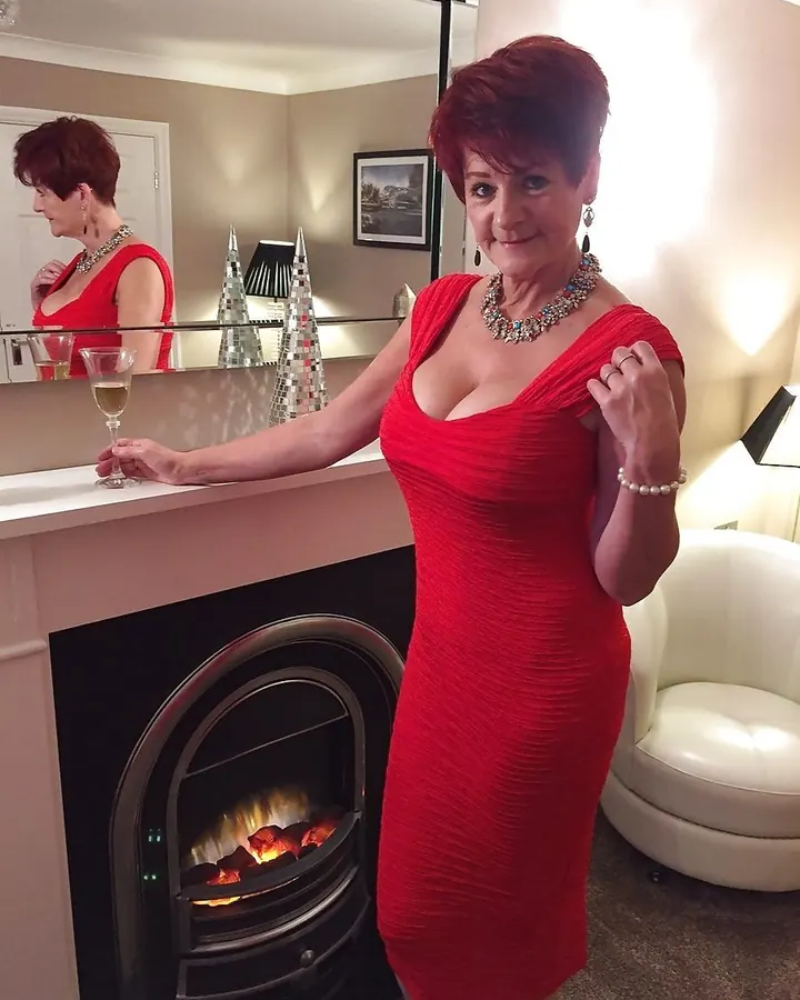Femme aux cheveux roux portant une robe rouge ajustée, debout près d'une cheminée allumée dans un salon élégant.