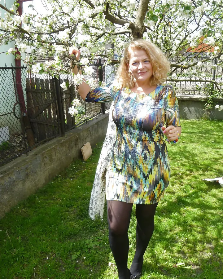 Femme aux cheveux blonds portant une robe colorée à motifs géométriques, debout sous un arbre en fleurs dans un jardin.