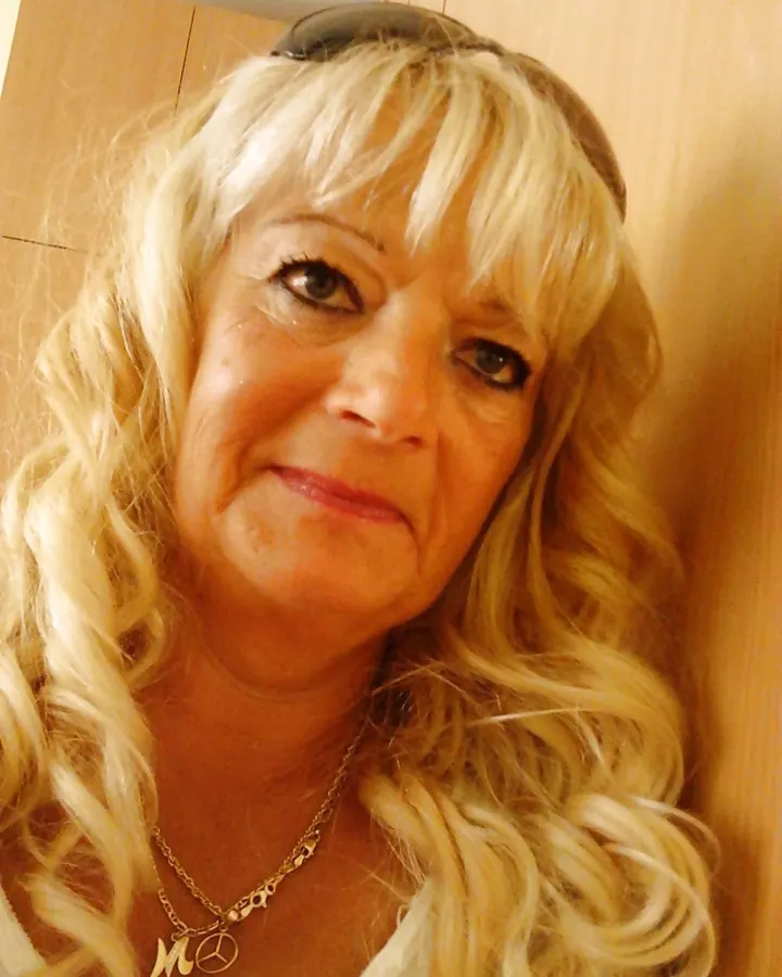 Femme blonde aux cheveux courts portant un pull bleu clair, assise sur un lit blanc avec un collier en forme de cœur, souriant à l'appareil.