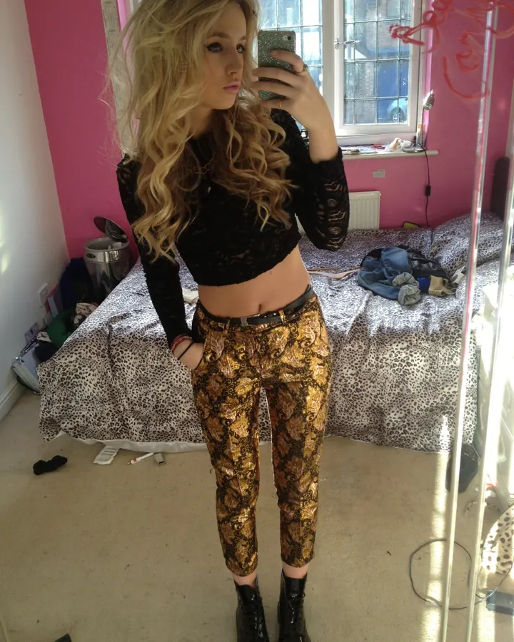 Femme blonde aux cheveux bouclés portant un haut noir en dentelle et un pantalon doré à motif, prenant un selfie devant un miroir dans une chambre rose.