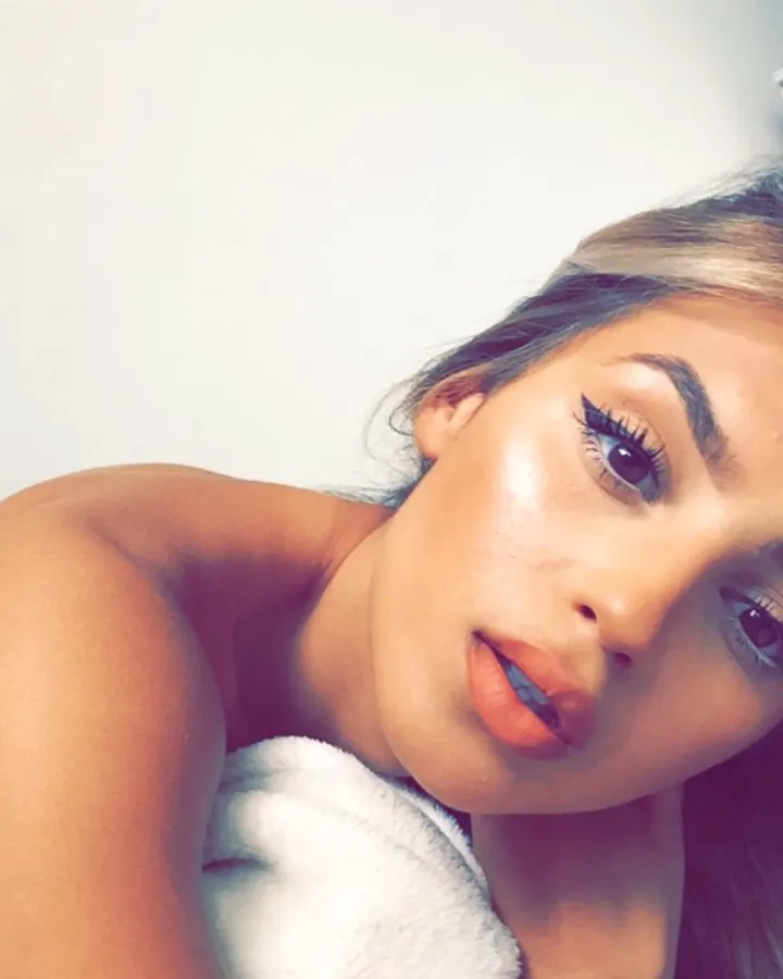 Femme blonde portant un haut noir et un pantalon doré, prenant un selfie devant un miroir dans une chambre rose.