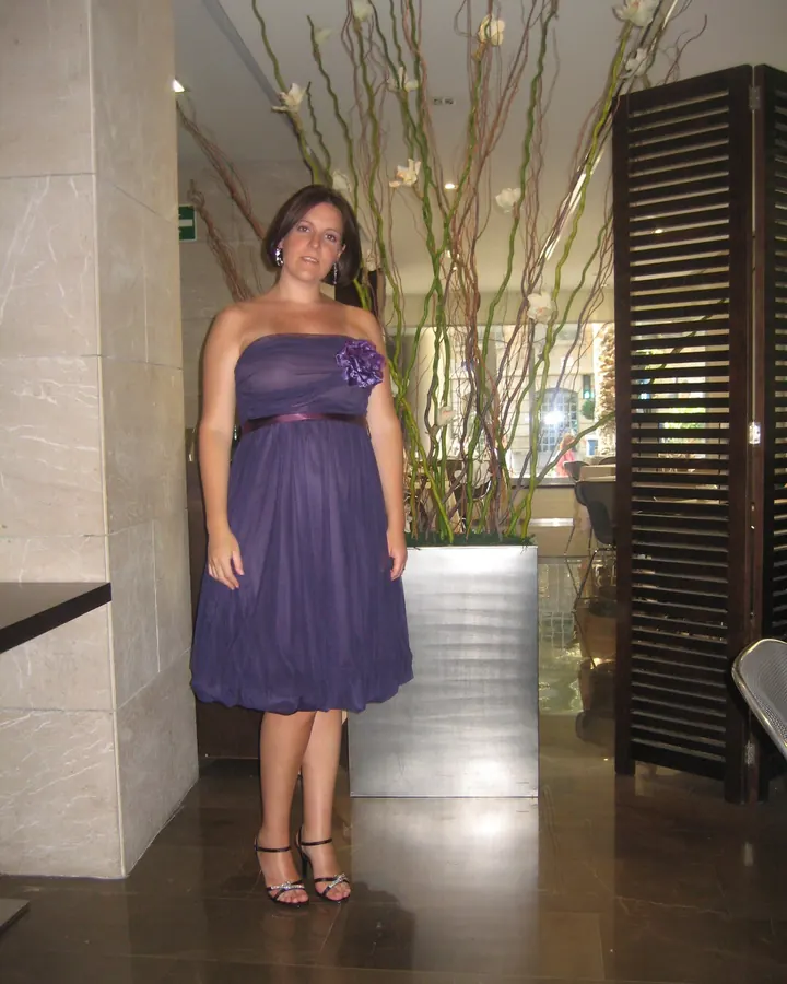 Femme en robe violette sans bretelles, debout dans un hall élégant avec un grand décor floral derrière elle.