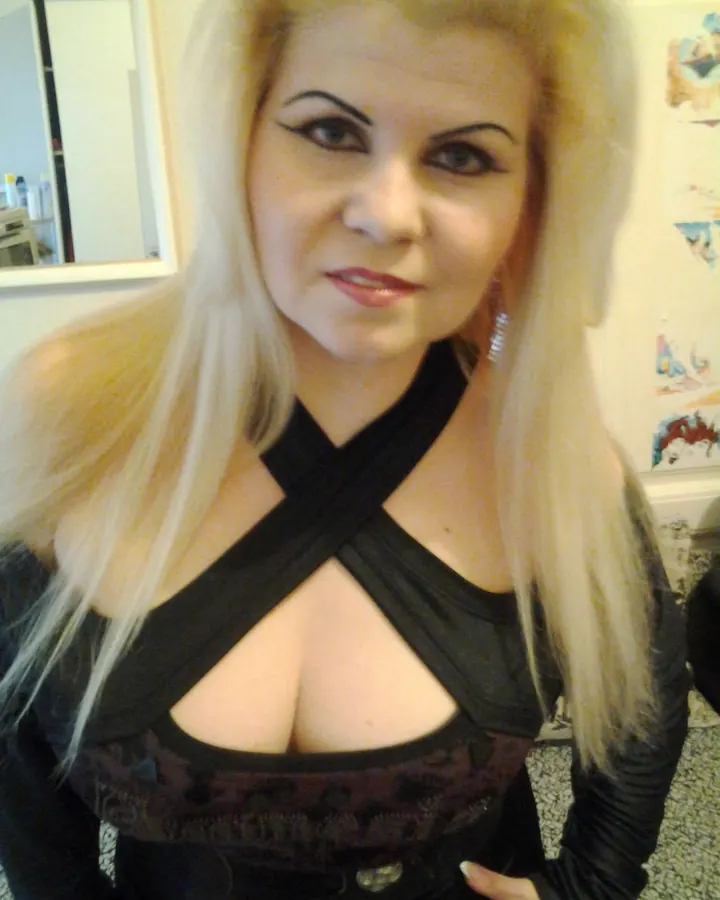 Femme blonde en haut noir à découpes croisées, regardant directement l'appareil, dans un intérieur domestique.