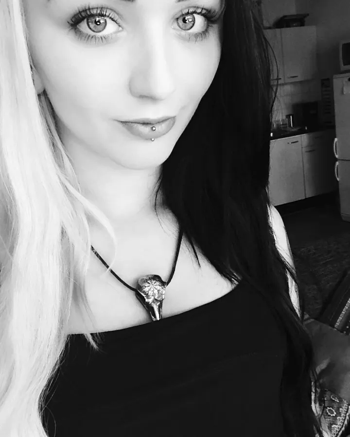 Portrait en noir et blanc d'une jeune femme aux yeux gris, cheveux blonds et noirs, piercing à la lèvre, portant un collier avec pendentif oiseau.