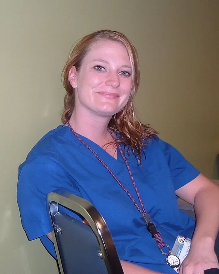 Femme aux cheveux blonds en tenue médicale bleue, assise sur une chaise dans une pièce avec un bureau en arrière-plan