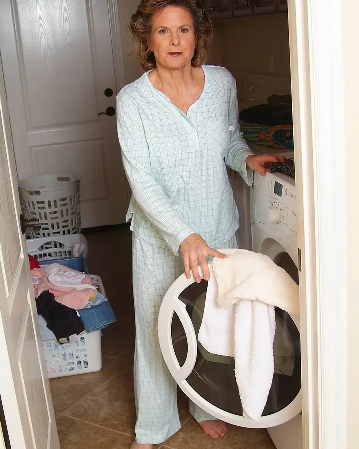 Femme aux cheveux bruns portant un pyjama à carreaux, debout devant une machine à laver dans une buanderie
