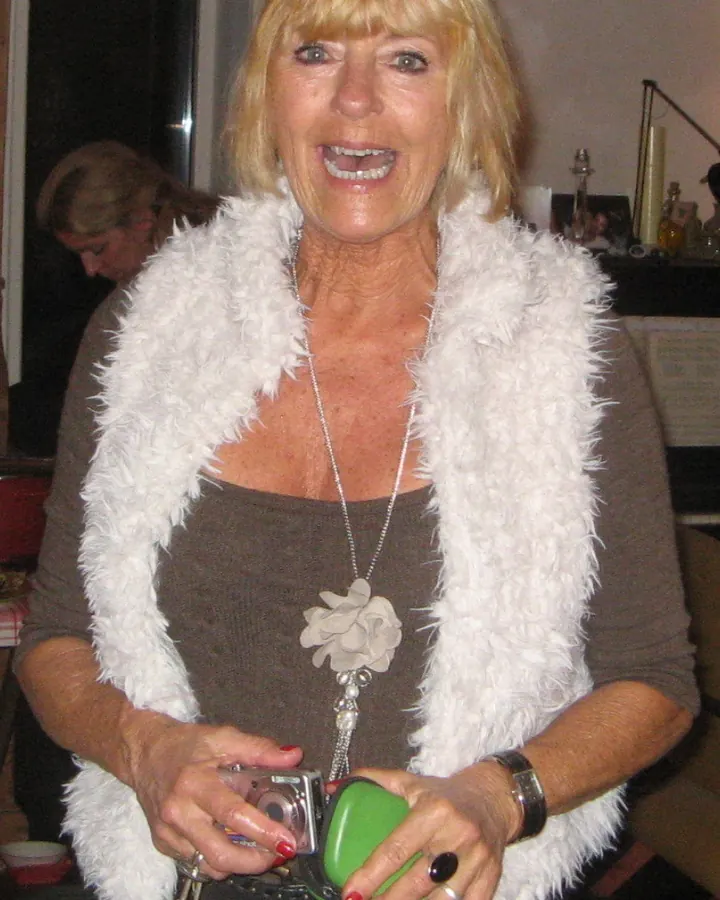 Femme blonde aux cheveux courts, en gilet blanc fourré, tenant un appareil photo et un sac vert.