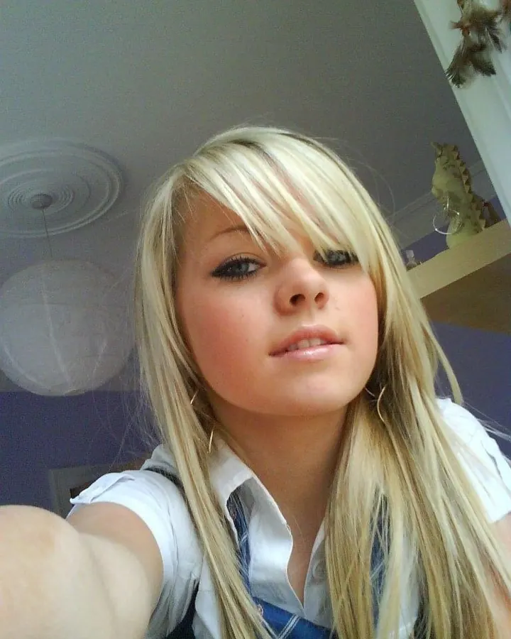 Femme blonde aux cheveux longs, en haut blanc et jupe bleue à motifs floraux, prenant un selfie dans une cabine d'essayage.