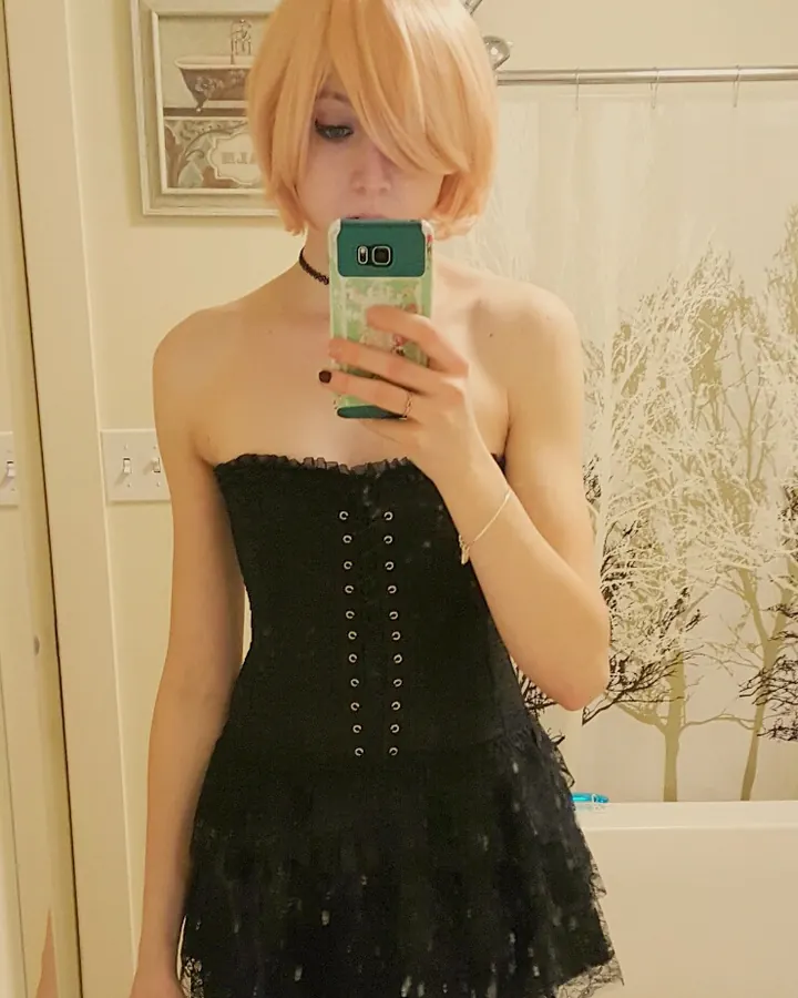 Femme blonde aux cheveux courts, en corset noir, prenant un selfie dans une salle de bain.