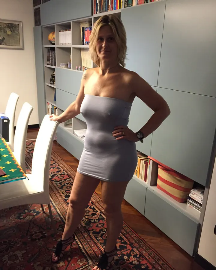 Femme blonde aux cheveux courts, en robe grise sans bretelles, posant dans une pièce avec bibliothèque.