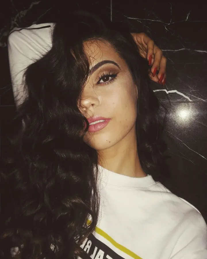 Femme aux cheveux noirs bouclés, maquillée, portant un t-shirt blanc, allongée sur un lit sombre.