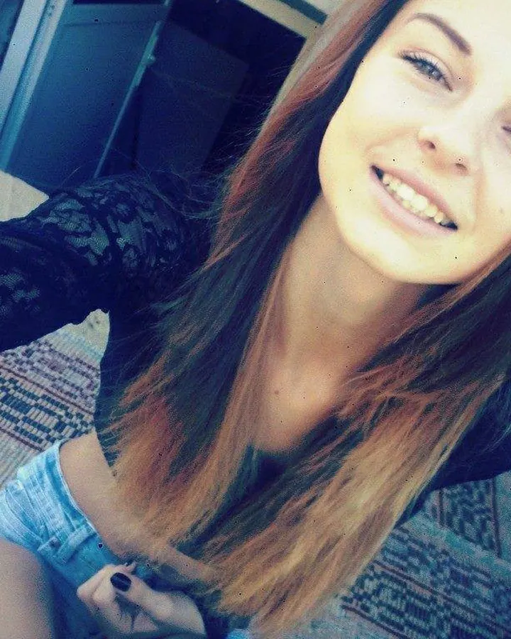 Femme aux cheveux châtains portant un haut blanc et une jupe courte, posant de profil, les bras croisés, souriante, dans un parc urbain.