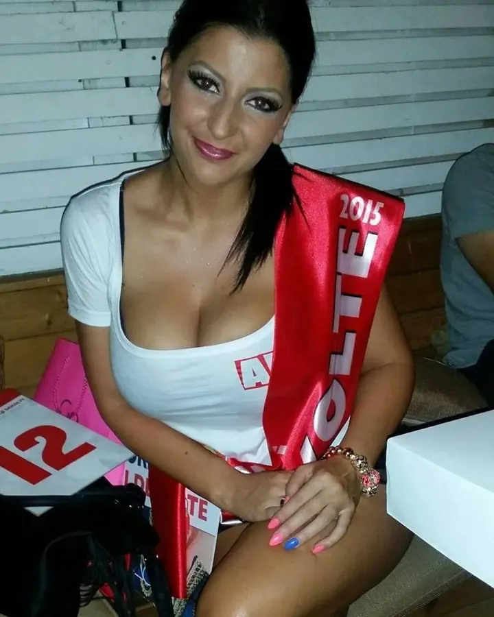 Femme aux cheveux bruns foncés assise, portant un t-shirt blanc et une écharpe rouge avec l'inscription '2015', souriant à la caméra, main posée sur la cuisse.