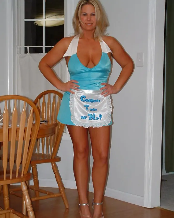 Femme blonde en robe turquoise avec tablier blanc portant l'inscription 'Coffee, Tea or Me?', pieds en sandales à talons, dans un cadre intérieur avec table en bois.