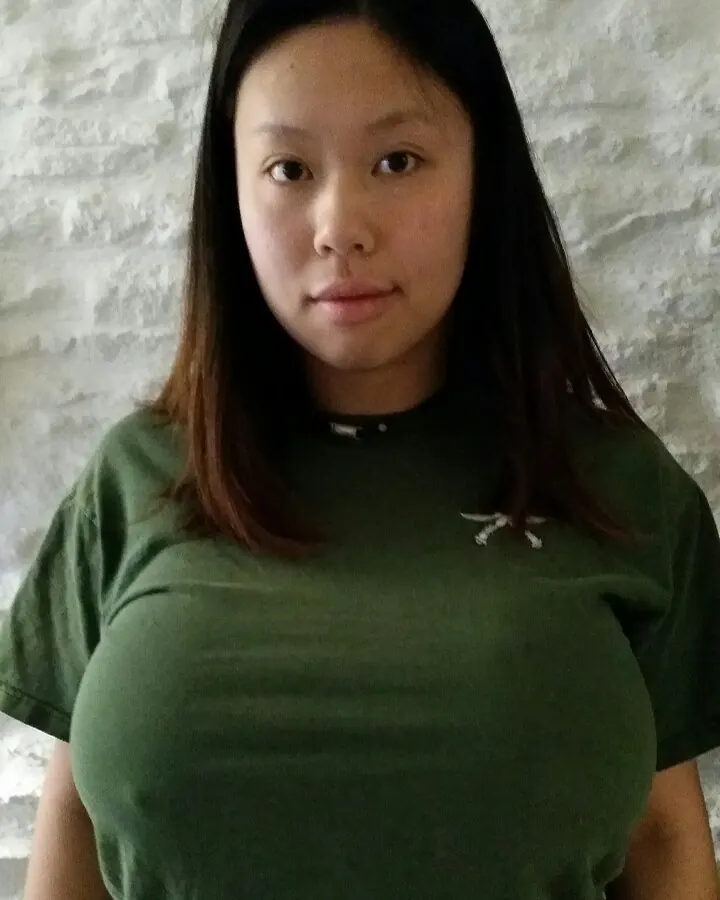 Femme aux cheveux noirs portant un t-shirt vert, regardant directement la caméra, debout devant un mur texturé blanc.