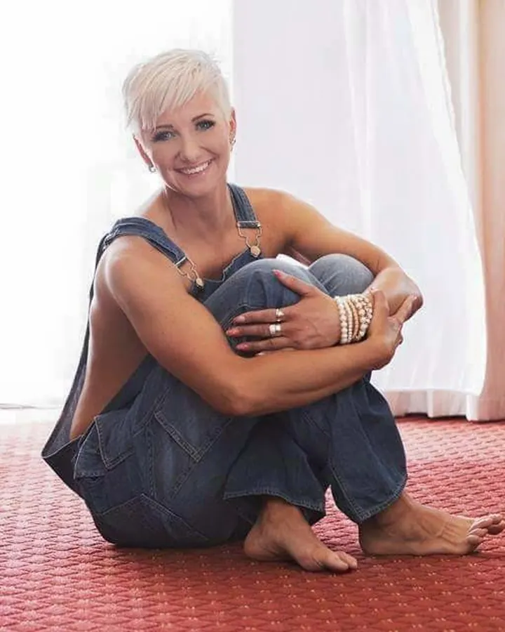 Femme aux cheveux blancs assise sur sol rouge, souriante, en salopette en jean