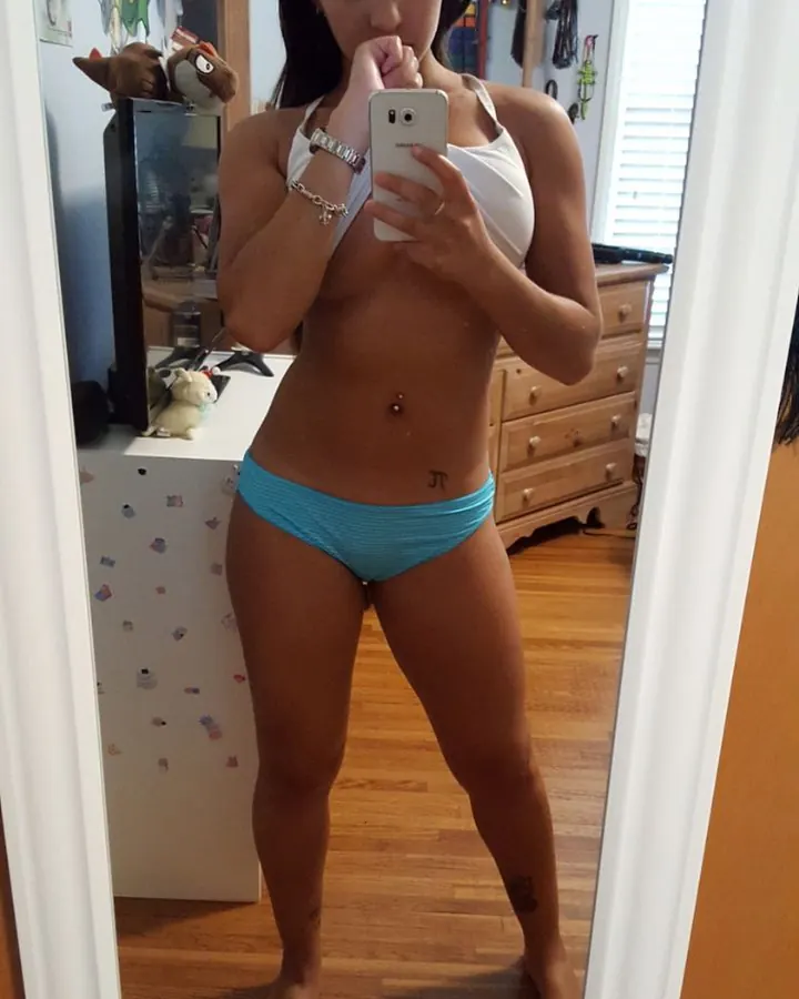 Femme aux cheveux foncés, prenant un selfie devant un miroir, portant un haut blanc et un short turquoise, dans une chambre.
