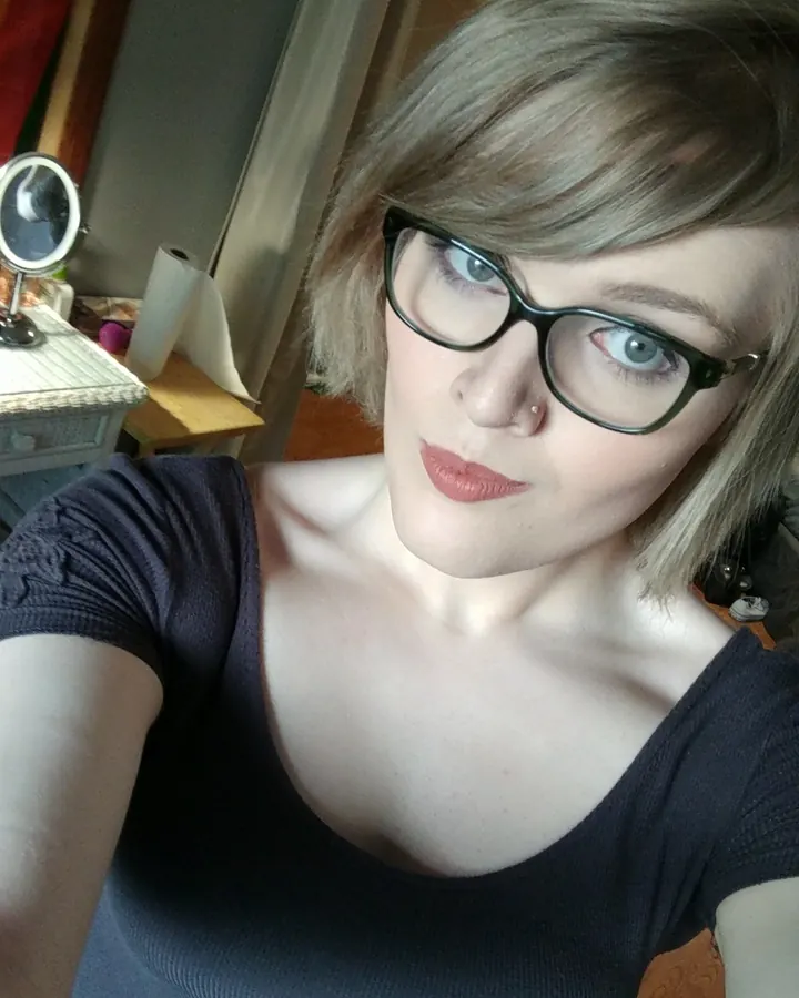 Femme aux cheveux blonds courts, portant des lunettes noires, un haut noir, et regardant l'appareil photo dans un cadre intérieur.