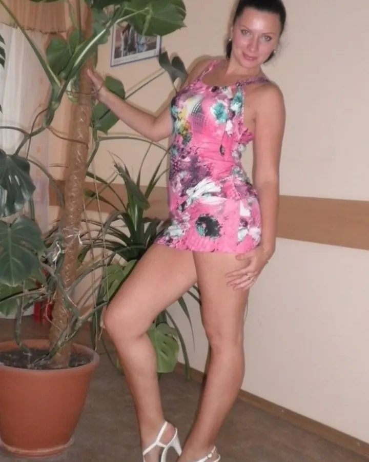 Femme en robe rose à motifs floraux, posant près d'une grande plante verte, chaussée de talons blancs.