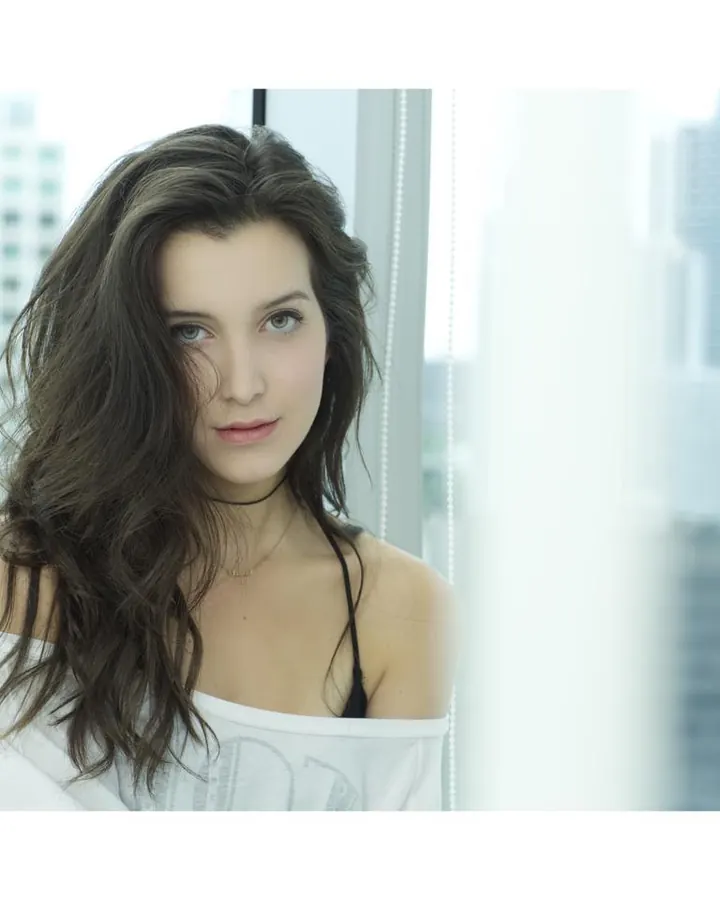 Femme brune aux yeux gris en pull blanc près d'une fenêtre