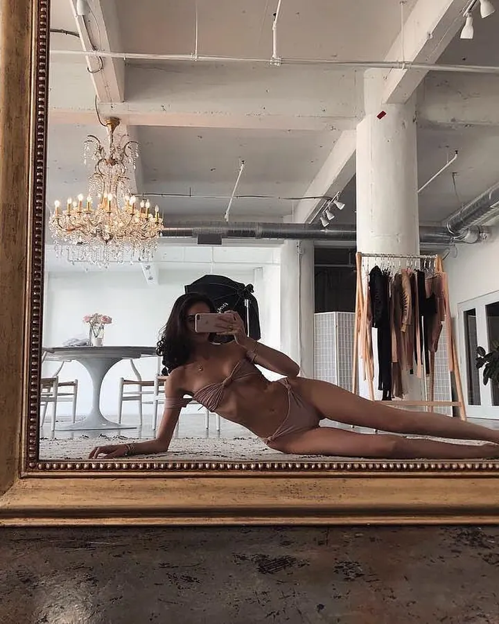 Jeune femme en lingerie beige, prenant un selfie dans un grand miroir doré dans un loft lumineux.