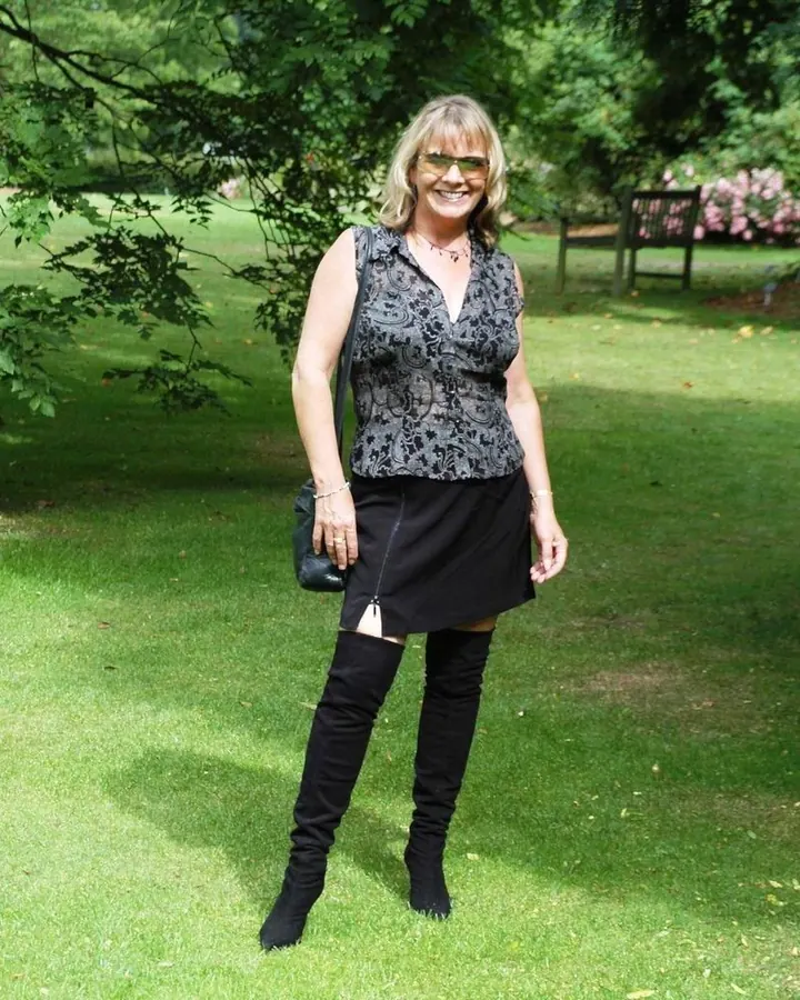 Femme blonde en haut noir à motifs et jupe noire, portant des bottes hautes, debout sur une pelouse verte.