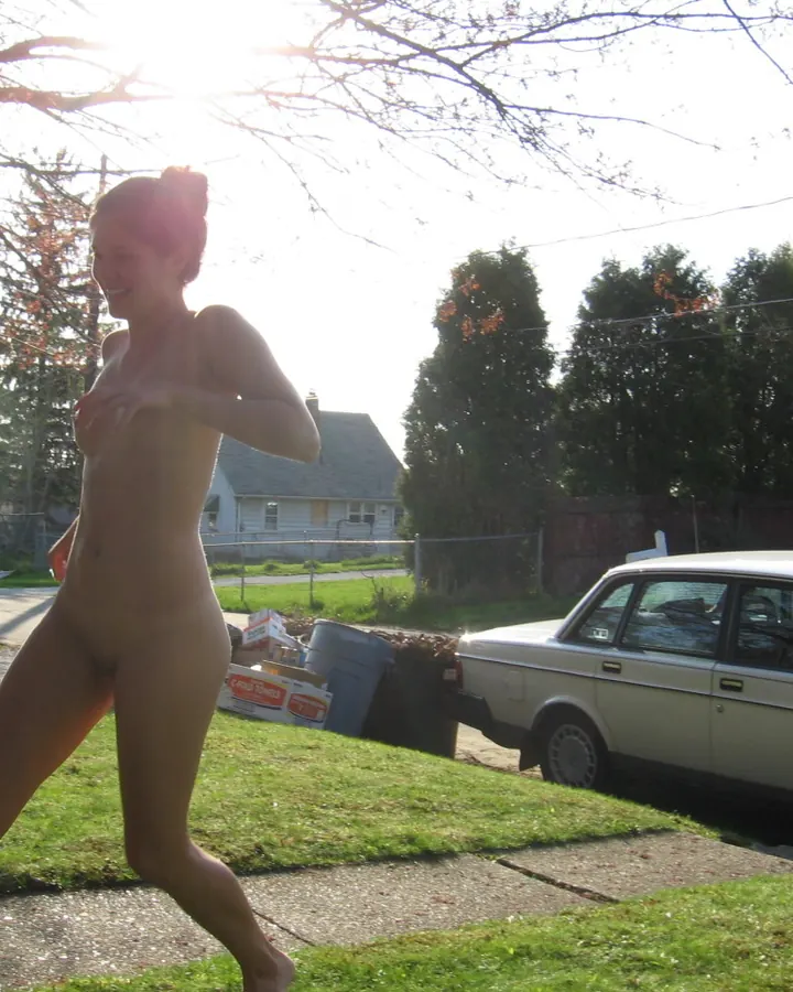 Femme nue, souriante, marchant sur une pelouse devant une maison, le soleil derrière elle.