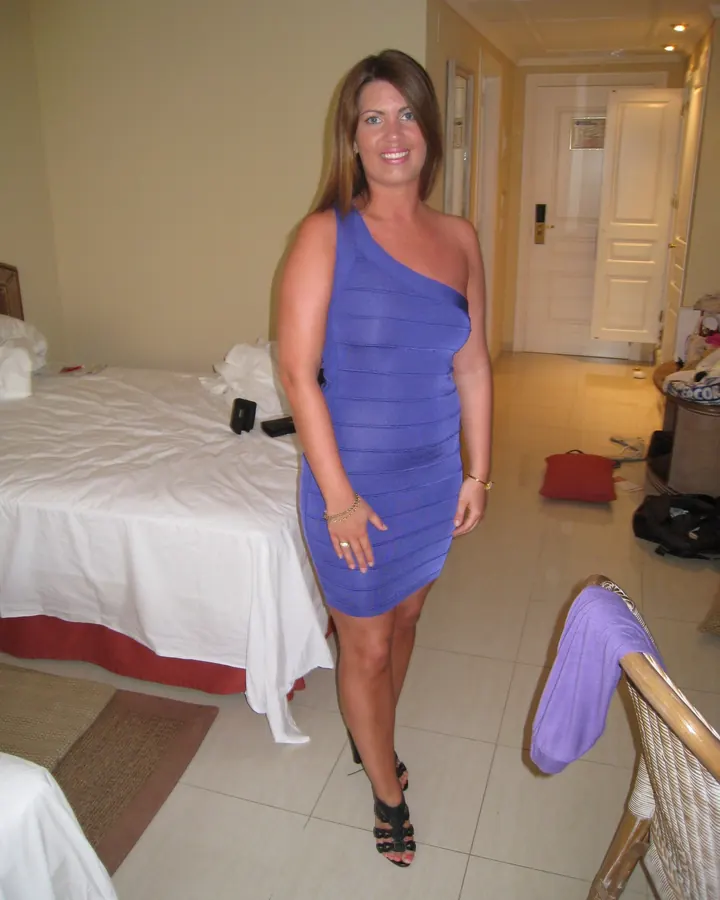 Femme en robe bleue asymétrique, souriante, dans une chambre d'hôtel avec un lit en arrière-plan.