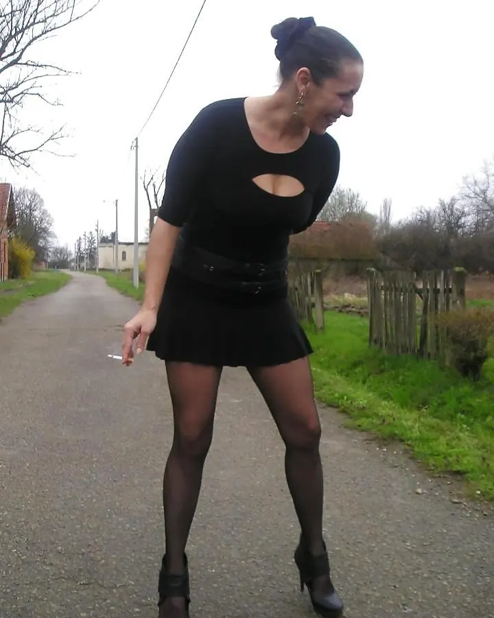 Femme en tenue noire, chemisier avec découpe, jupe courte, collants et bottines, fumant une cigarette sur un chemin rural.