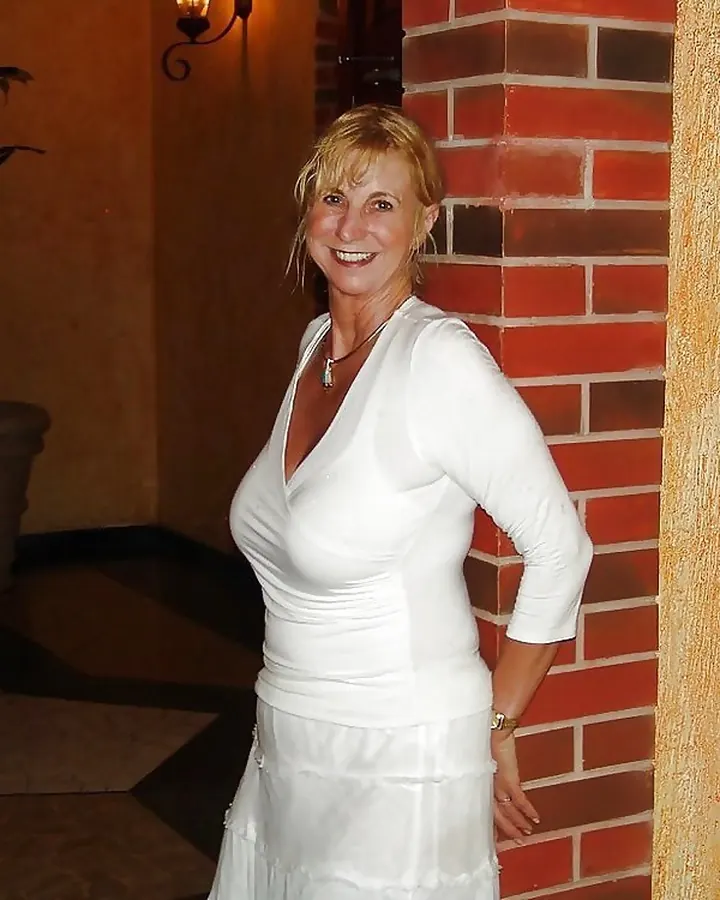 Femme en robe blanche, souriant, appuyée contre un mur en briques