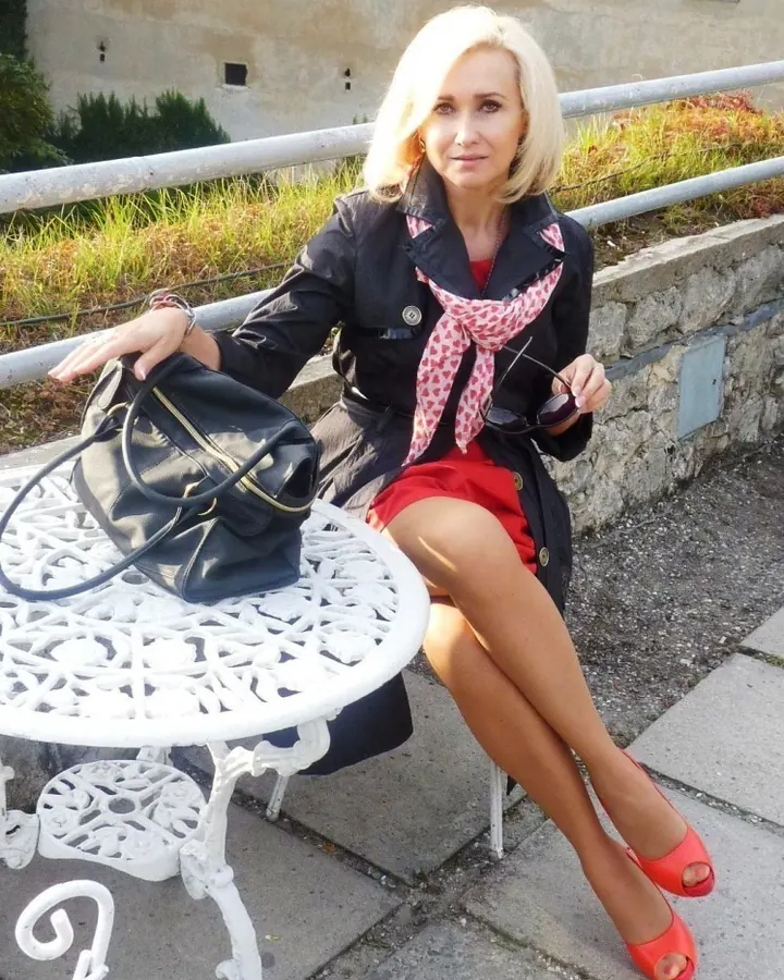 Femme blonde en chemise bleue à motifs, souriante devant un mur en bois, avec des boucles d'oreilles dorées.